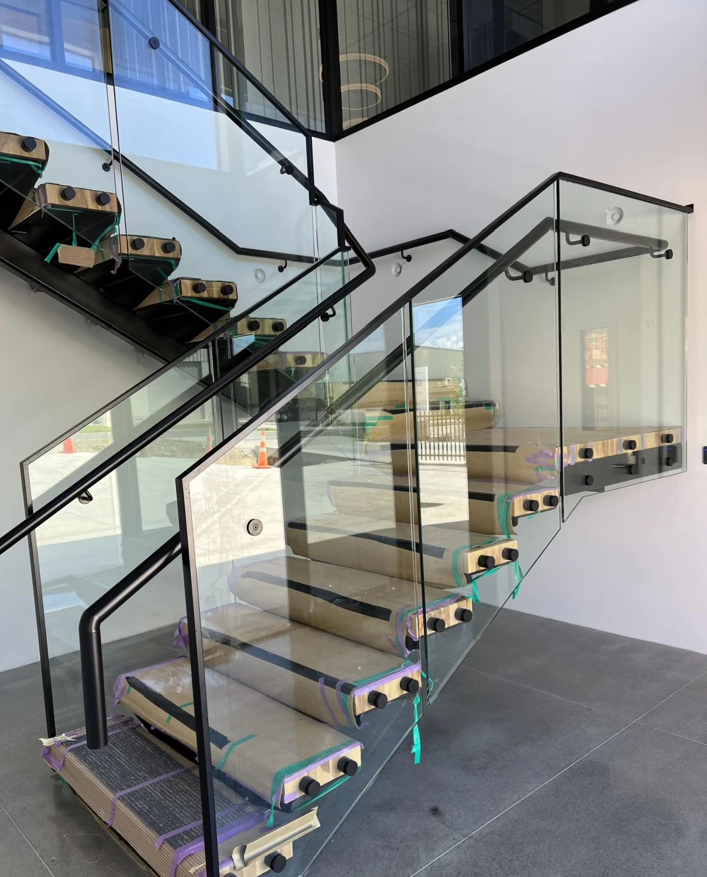 Glass stair panels with round side fixings and black handrails beside protected treads during installation