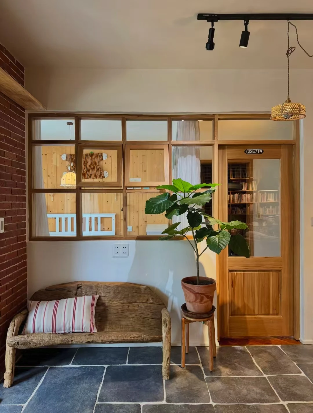 Wood-tone interior window with divided panes beside a matching glazed door, wicker bench and potted plant