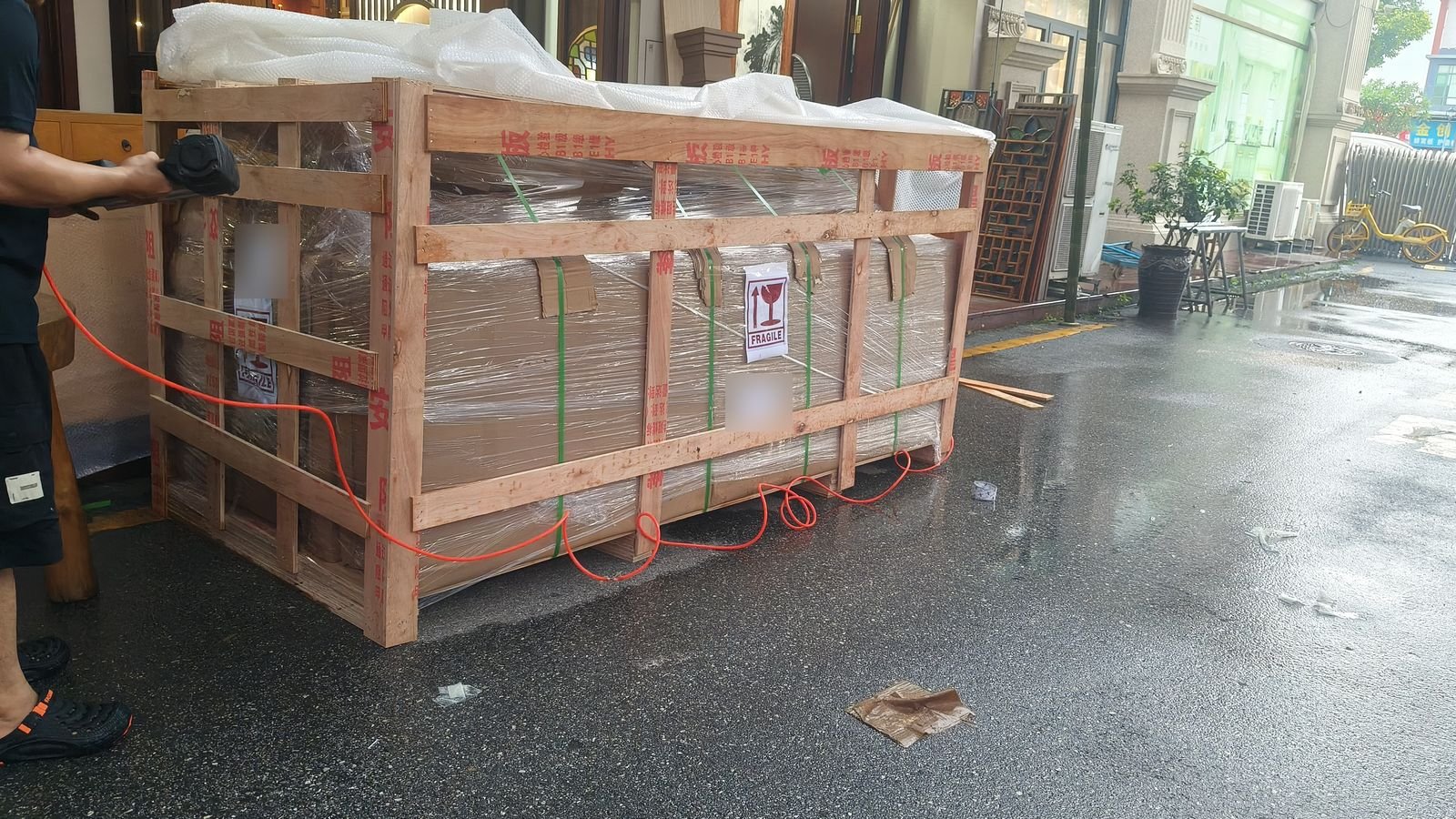 Worker building a wooden crate around stretch-wrapped cartons with green strapping and a FRAGILE label
